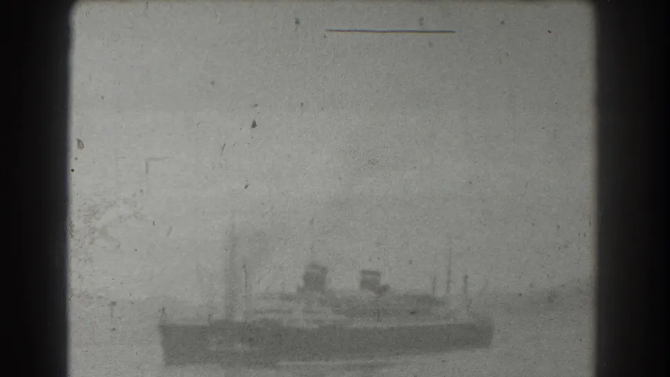 Distant 1939 home-movie view of a large vessel in a bay, with funnels and rigging softened by haze and film grain.