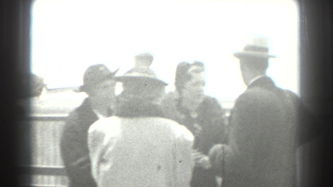 Adults in hats and coats gather near an airfield railing in bright backlight.