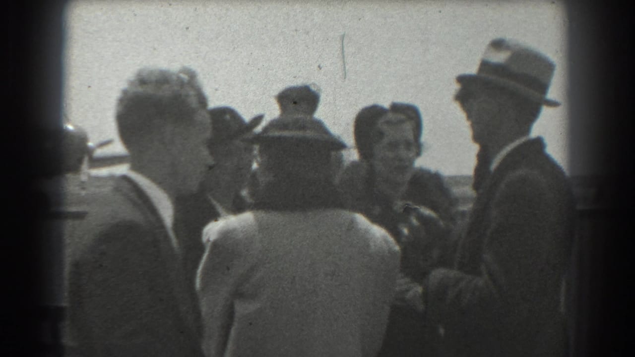 Group of adults in suits and hats converse beside an airfield area.