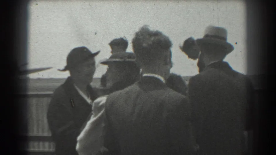 1939 San Francisco airbase frame with adults in formal clothing gathered near a railing, seen from a handheld home-movie viewpoint.