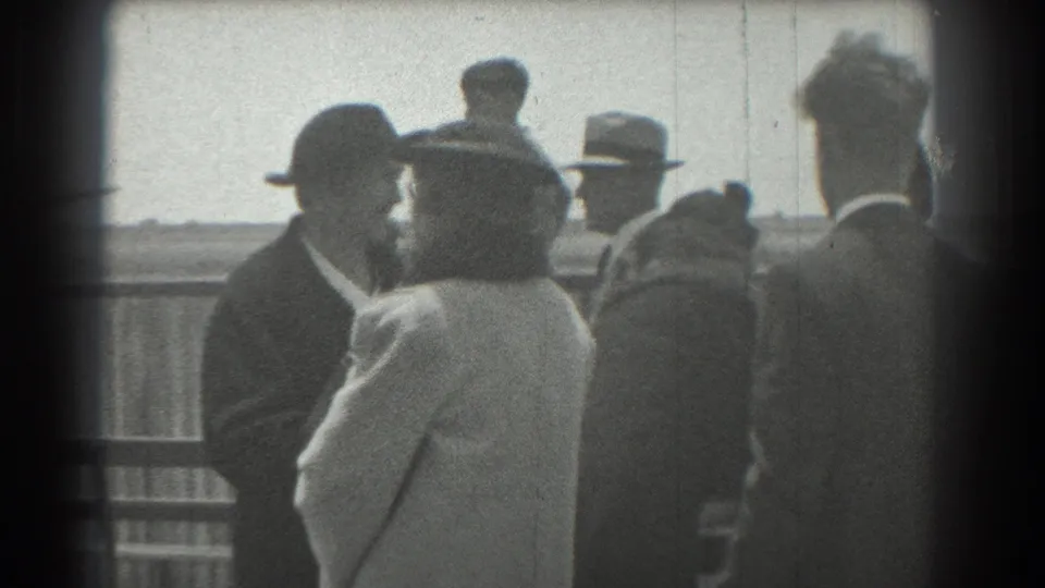 Adults in coats and brimmed hats stand in conversation near a railing, with open landscape or water beyond in 1939 San Francisco footage.