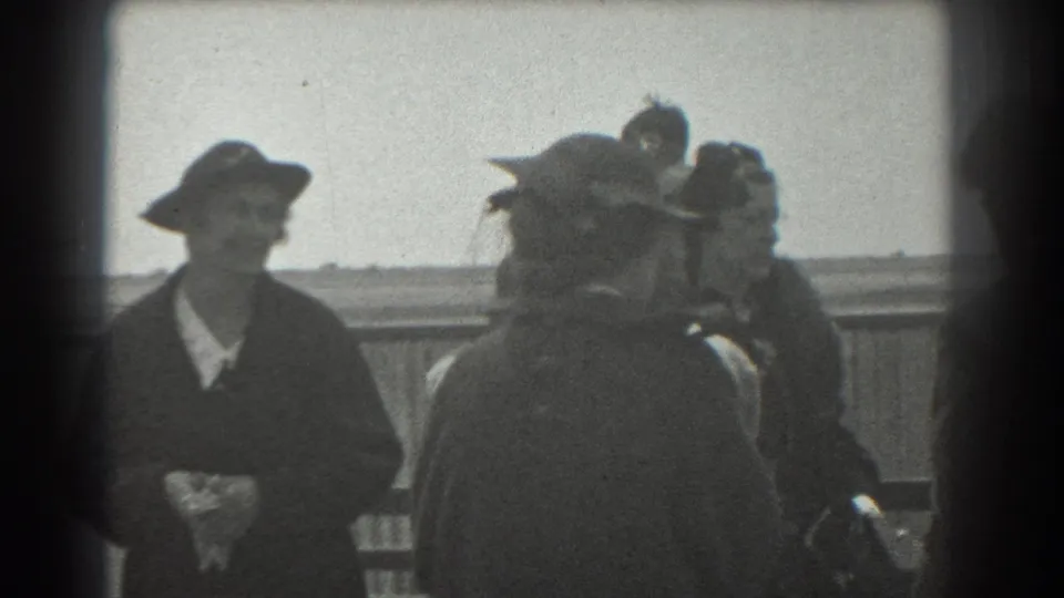 1939 San Francisco frame of a woman in a wide-brimmed hat and dark coat among adults on an outdoor platform.