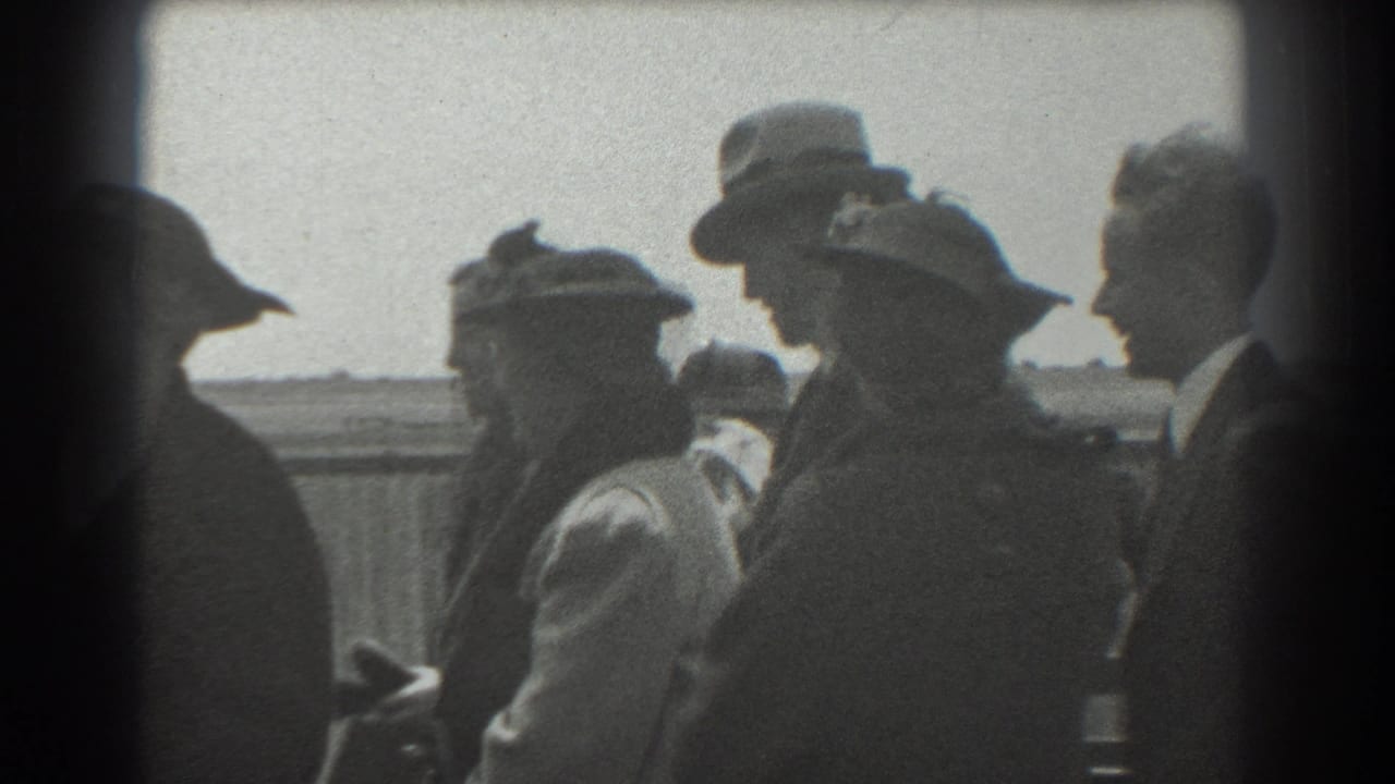 Adults in hats and coats stand near a fence in bright daylight.