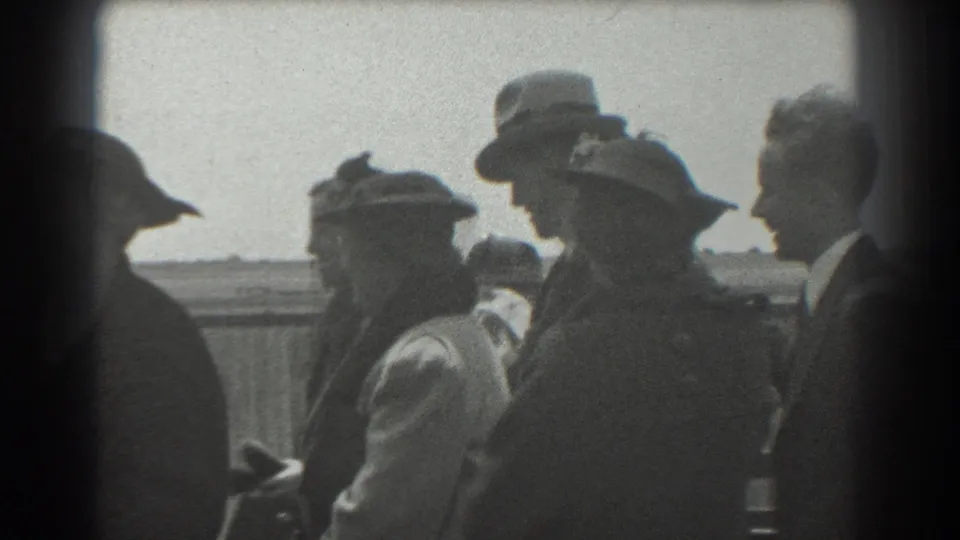 1939 San Francisco home-movie frame showing adults in hats and coats gathered near a fence under bright sky.