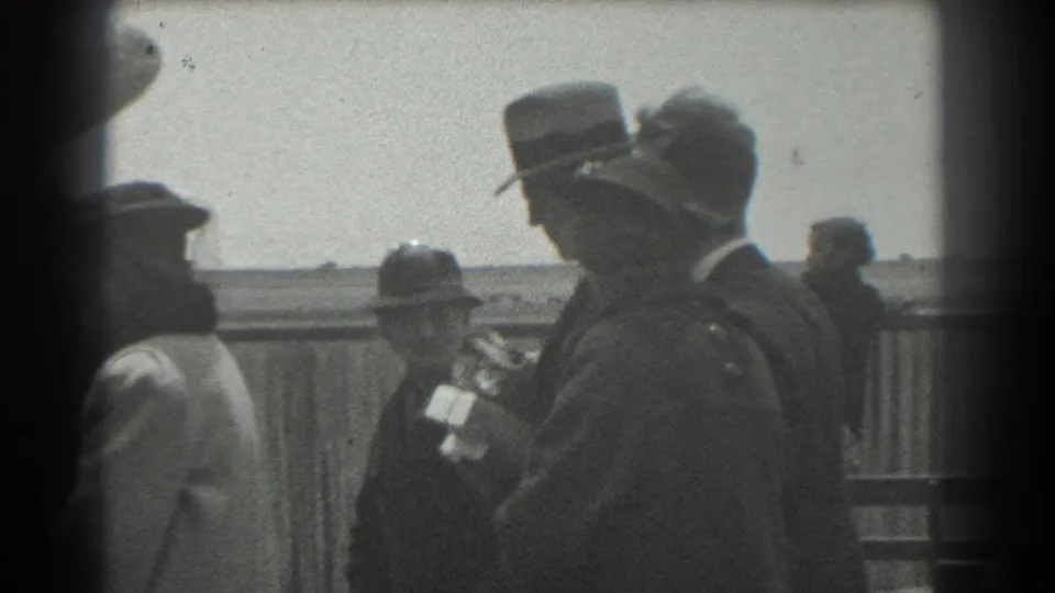 San Francisco 1939 home-movie frame with adults in hats moving past a fence, including one person carrying pale papers or packages.