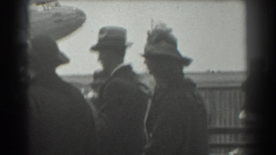 In 1939 San Francisco airport footage, suited adults and hat-wearing spectators stand near a barrier with a plane partly visible behind them.