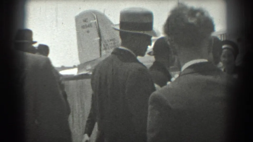 At the San Francisco airport, suited spectators cluster near a United Airlines aircraft tail in grainy black-and-white 8mm footage.
