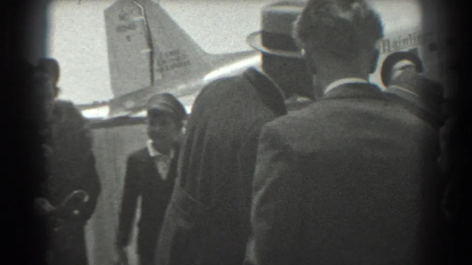 The 1939 San Francisco airport crowd remains gathered near a United Airlines aircraft, framed through the backs of suited spectators.