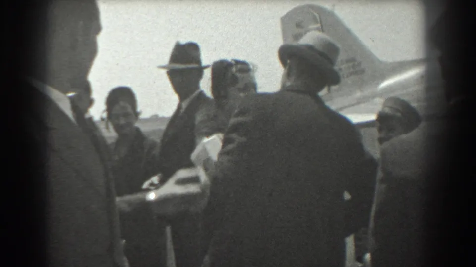 1939 San Francisco airport home-movie frame with suited men, women in hats, and an aircraft tail behind the group.