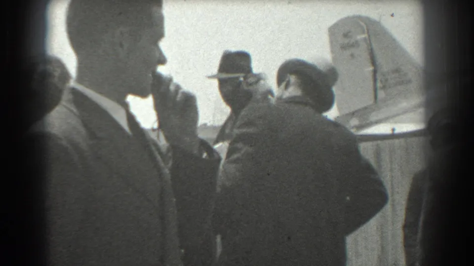 Close airport-side view of a suited man smoking, with formally dressed greeters and an aircraft tail in the background.