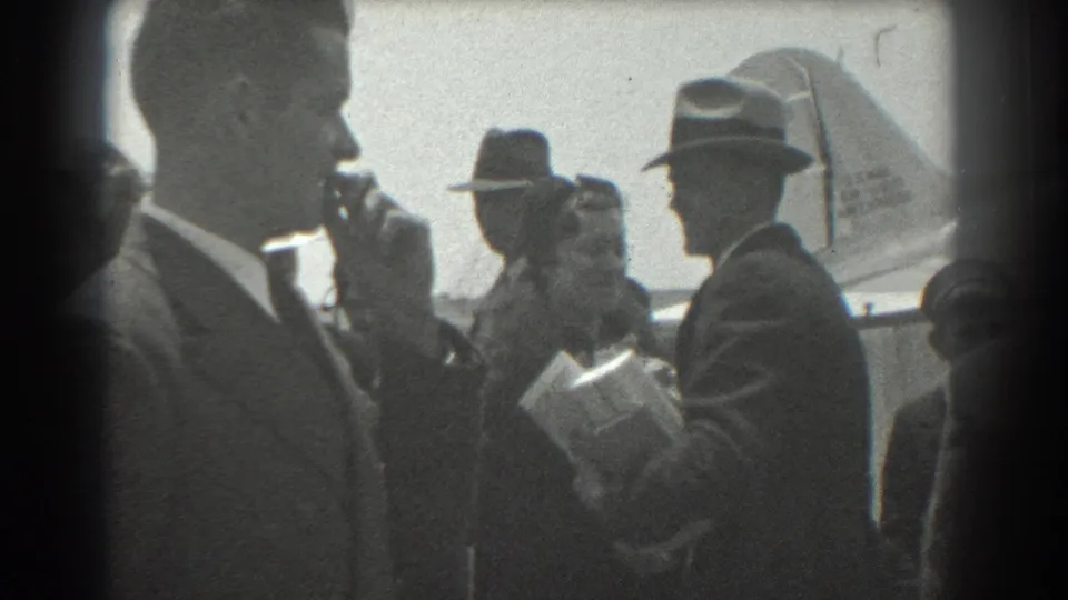 1939 San Francisco airport frame showing adults in formal clothing, one holding papers or packages, with a plane tail behind them.