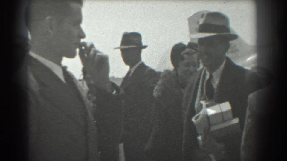 Close 1939 San Francisco airport view with a man carrying wrapped gifts, a suited smoker, and other greeters near the aircraft.