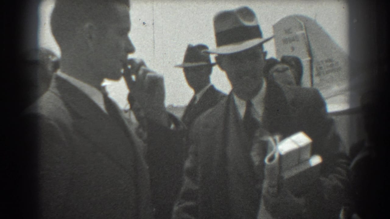 In 1939 San Francisco airport footage, men in suits and brimmed hats stand near an aircraft as one man holds papers or packages and another appears to greet him.