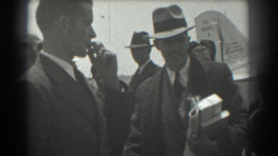 In 1939 San Francisco airport footage, men in suits and brimmed hats stand near an aircraft as one man holds papers or packages and another appears to greet him.