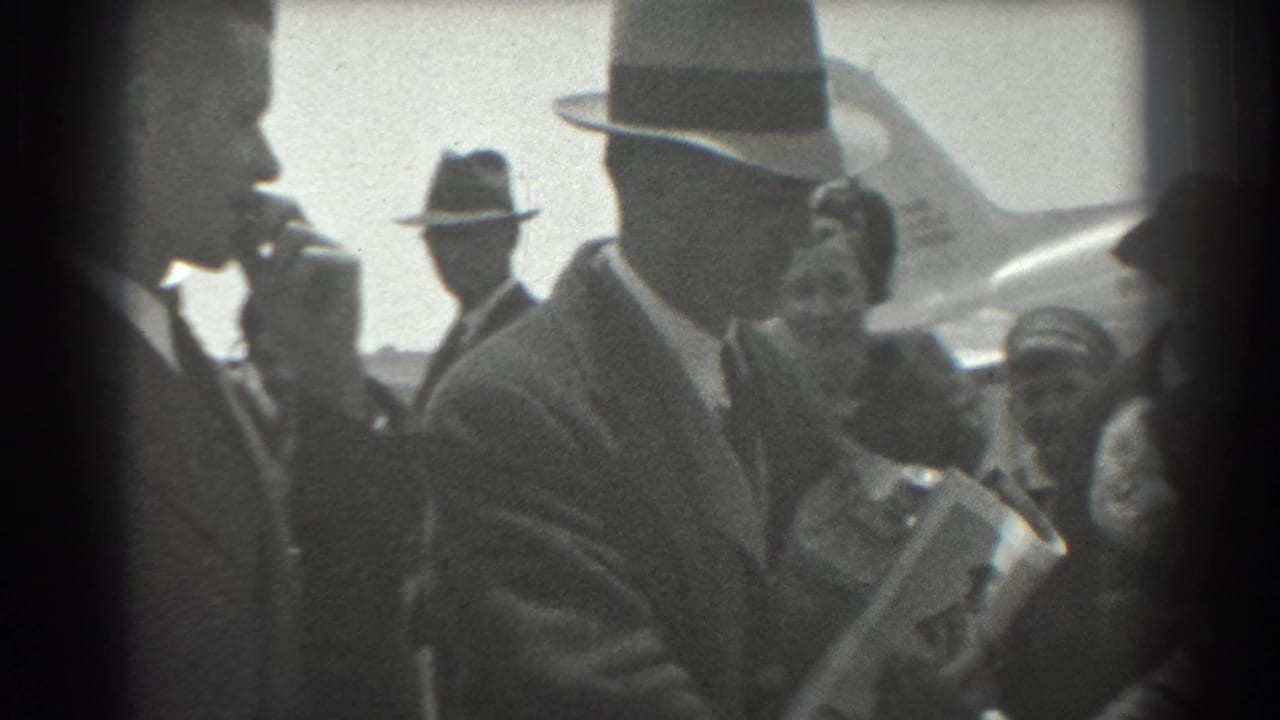 A man in a fedora carries papers or packages beside other people near an airplane.