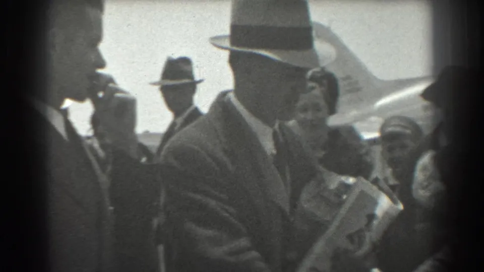 In 1939 San Francisco airport footage, a fedora-wearing man carries papers or packages while other adults stand close to an airplane in the background.
