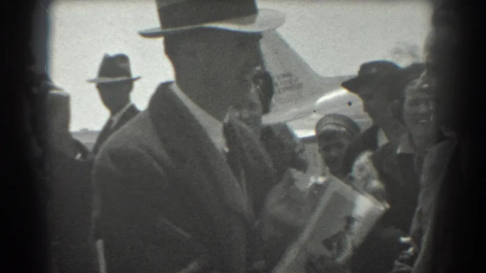 In 1939 San Francisco airport footage, a suited man in a fedora stands near smiling bystanders with an airplane visible behind the group.