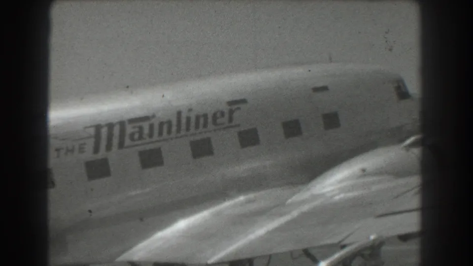 1939 San Francisco frame showing The Mainliner fuselage lettering, cabin windows, wing, and propeller aircraft nose area in monochrome 8mm footage.