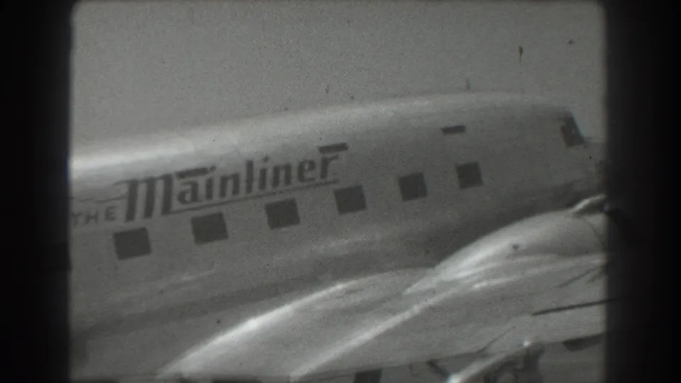 Black-and-white 8mm frame of The Mainliner aircraft side, with fuselage lettering, passenger windows, and a nearby wing surface.