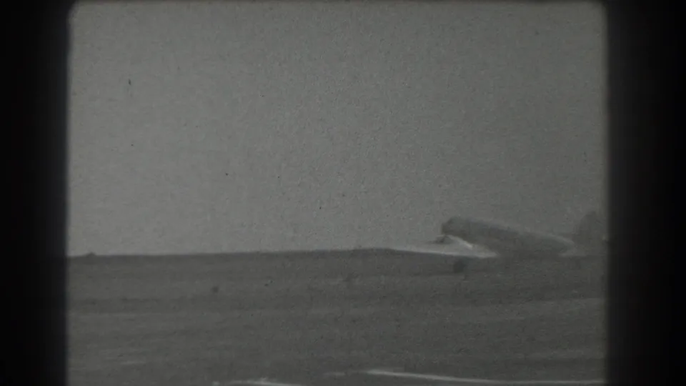 A small aircraft rolls across the open airfield in the lower frame, captured in soft black-and-white 8mm texture.