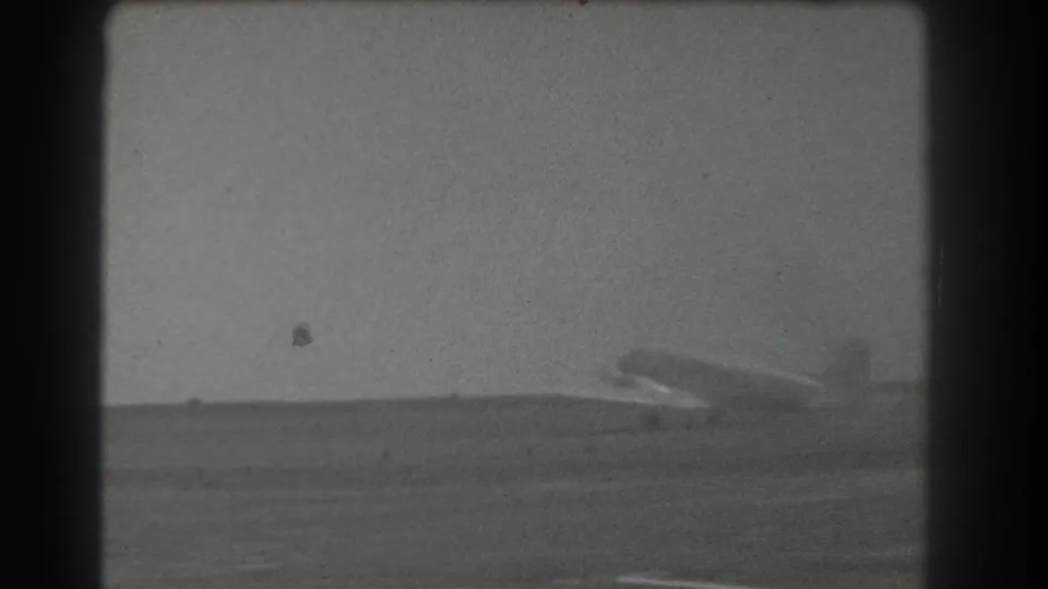 Black-and-white 8mm frame of a light propeller airplane on a runway near San Francisco, seen from a fixed distance.