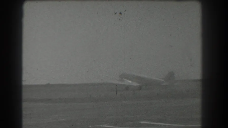 1939 San Francisco home-movie frame showing a distant airplane across an open airfield in soft black-and-white focus.