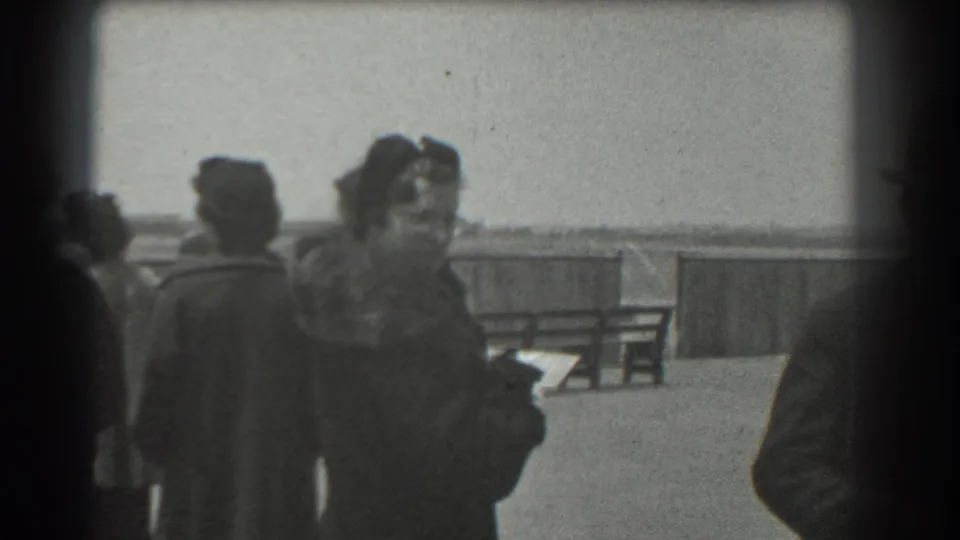 At a 1939 San Francisco airfield, a woman in a fur-trimmed coat turns while other spectators stand nearby.