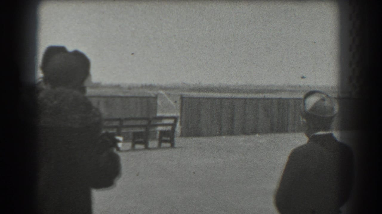 Women in dark coats and hats stand near a waterfront railing, seen from behind.