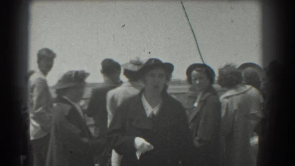 1939 San Francisco frame with adults in period dress near a railing, including a woman in a dark hat looking toward the camera.