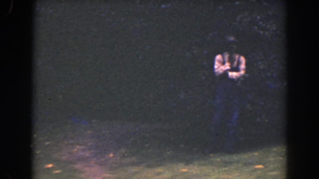 A small figure in a light shirt stands in a dim Florida outdoor scene, framed by heavy grain, dark foliage, and 8mm film borders.