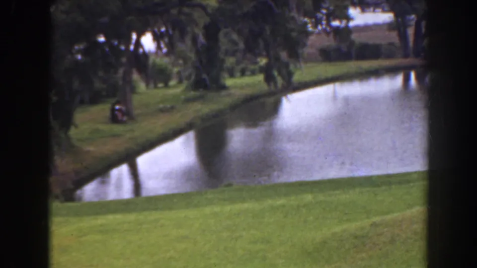 1941 Florida 8mm view of a golf-course pond bordered by green fairway and trees, seen through dark framing with a distant person at left.