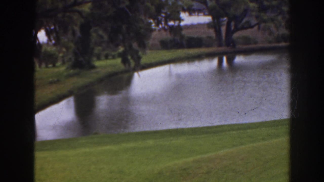1941 Florida home-movie frame showing a calm golf-course pond, bright grass, and trees viewed through dark vertical framing.