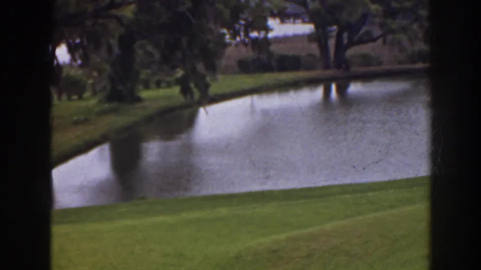 1941 Florida home-movie frame showing a calm golf-course pond, bright grass, and trees viewed through dark vertical framing.