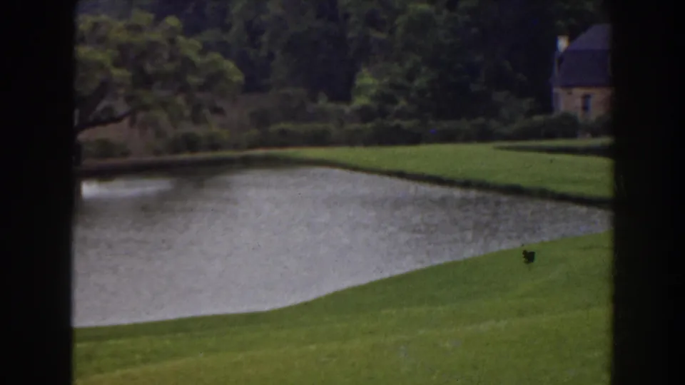 1941 Florida frame with a landscaped pond and a small stone house emerging beyond the lawn and trees.