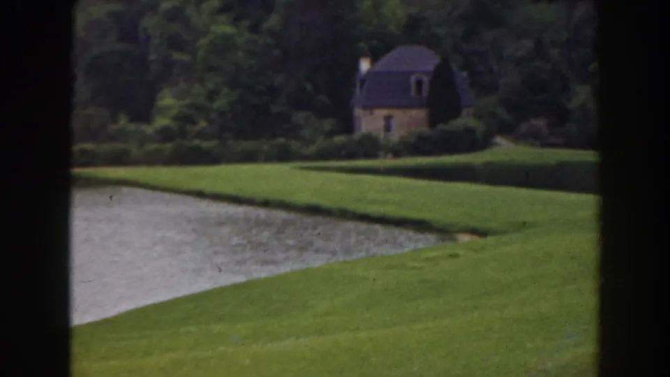 1941 Florida home-movie still with a grassy pond bank and a small stone house beyond trees and shrubs.