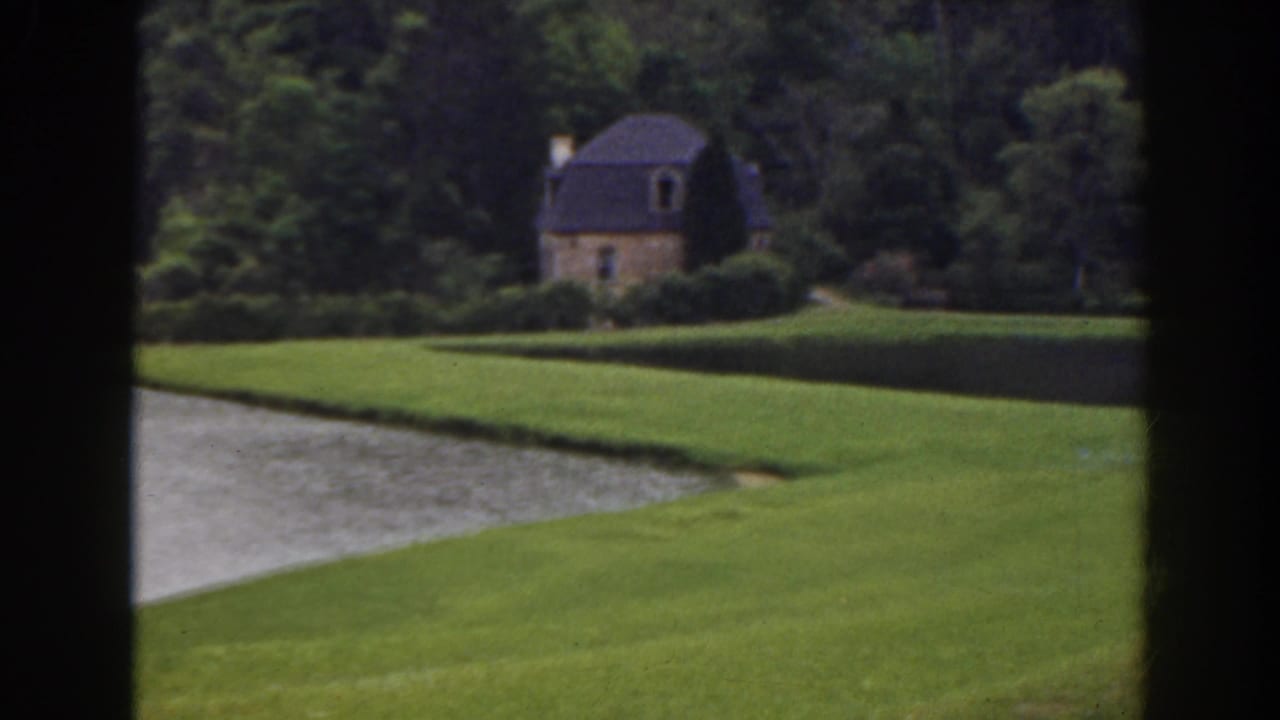 1941 Florida 8mm frame showing manicured grass, water, dense trees, and a distant cottage seen through dark window mullions.