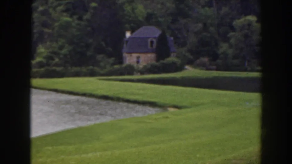 1941 Florida 8mm frame showing manicured grass, water, dense trees, and a distant cottage seen through dark window mullions.