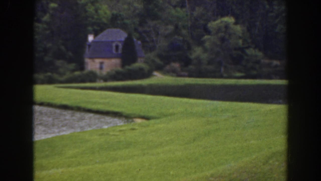 Manicured green lawn and water with a small cottage beyond trees, divided by a dark vertical frame.