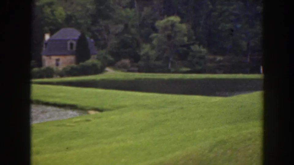1941 Florida 8mm frame with bright grass in the foreground, a pond edge, dense trees, and a small cottage near the left side.