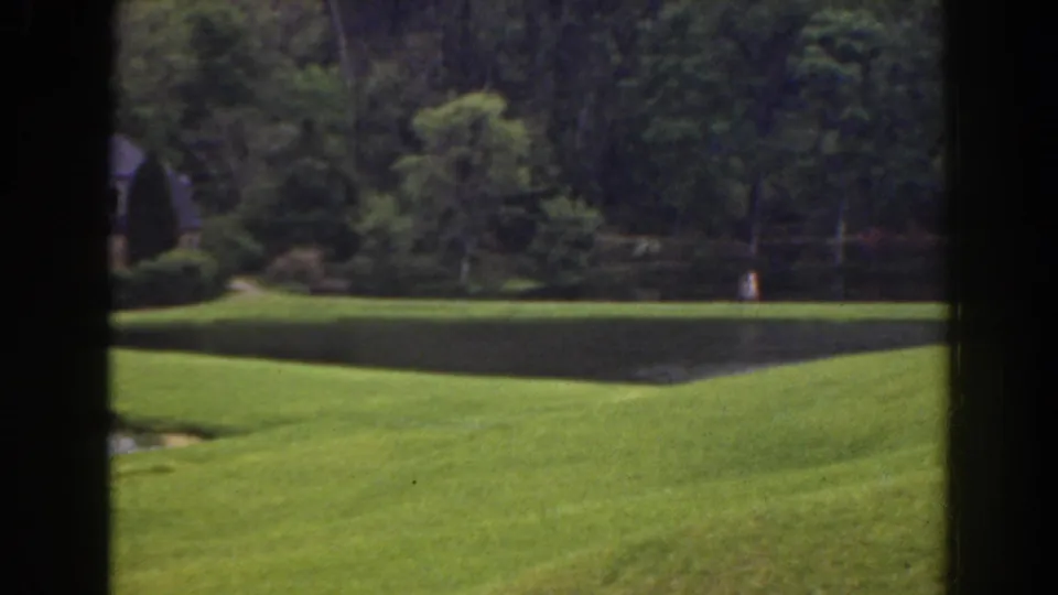 1941 Florida frame showing a manicured waterfront lawn, wooded backdrop, partly obscured cottage, and tiny distant people near the far bank.