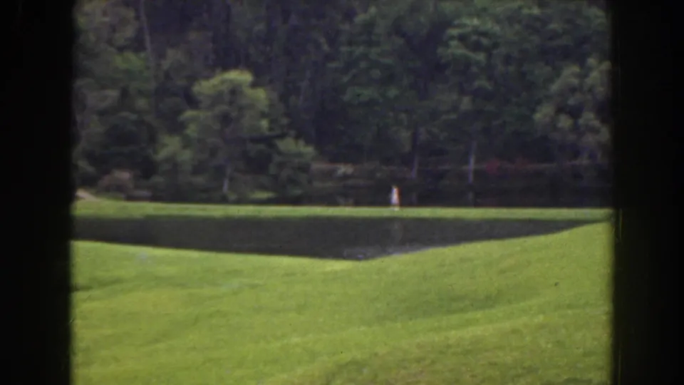 1941 Florida 8mm frame showing a distant person in white beside a pond, with rolling grass and dense trees behind.