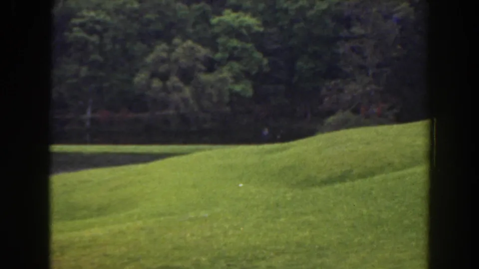 1941 Florida home-movie frame of a sloped lawn and pond with a barely visible figure near the tree-lined water.