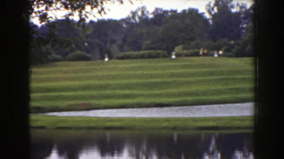 1941 Florida park landscape with distant people crossing a rolling lawn near shrubs, pond water, and a pale shoreline path.