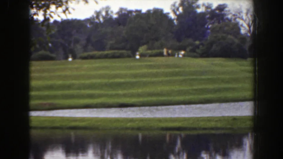 1941 Florida home-movie frame showing small distant figures on a park lawn above reflective pond water and a wooded backdrop.