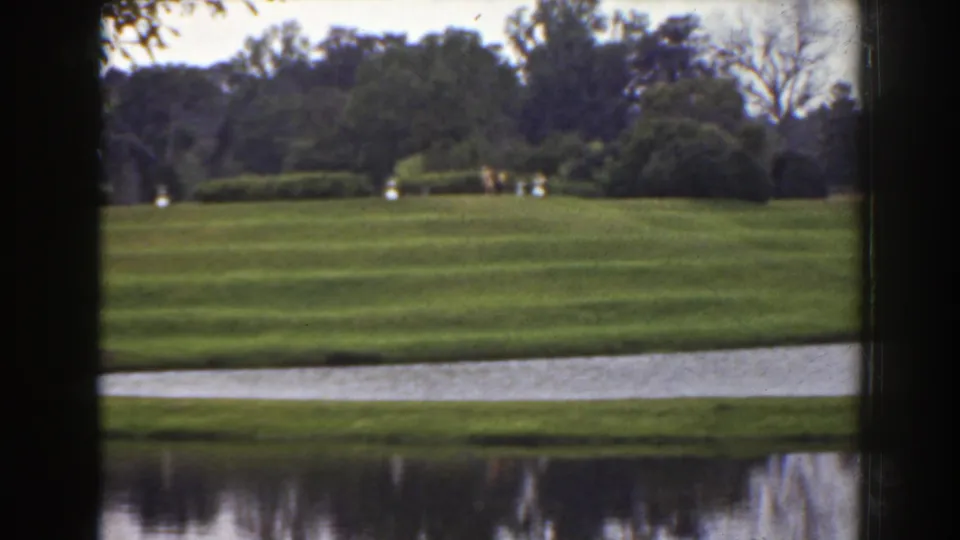 1941 Florida 8mm frame showing a striped green lawn, water in the foreground, trees beyond, and tiny distant figures in light clothing.