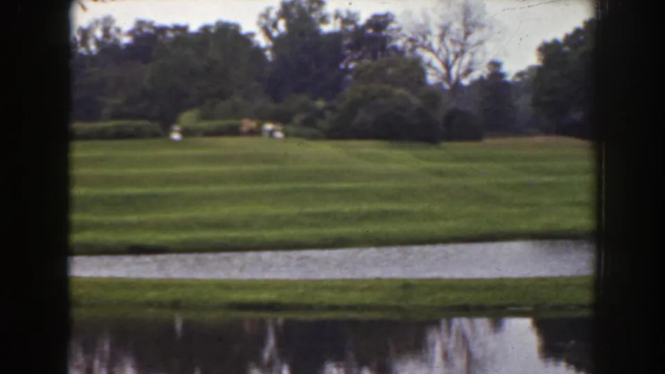 1941 Florida home-movie still of manicured grass beside a narrow water body, with hedges, trees, and small distant people crossing the lawn.