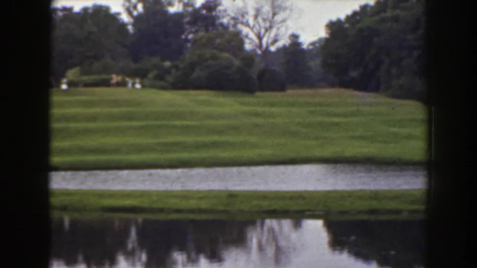 1941 Florida 8mm frame during the pan, showing a broad manicured lawn, water in the foreground, shrubs, and dense trees at the right edge.