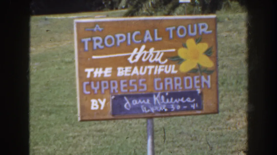 1941 Florida home-movie frame showing a hand-painted sign for a tropical tour through Cypress Garden in a grassy area.