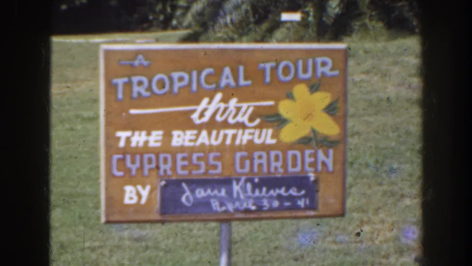 Close view of the Cypress Garden tropical tour sign, with painted lettering, a yellow flower, and a handwritten date panel.