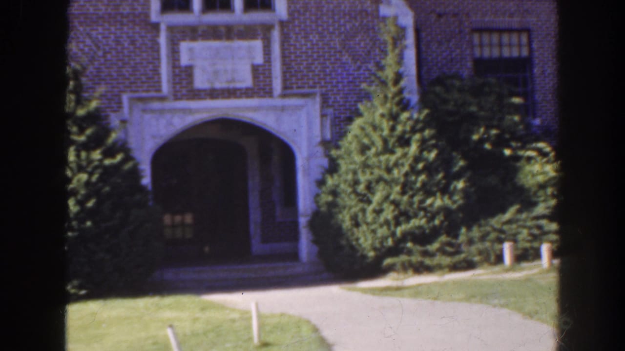 Brick building entrance with an arched doorway, shrubs, lawn, and curved paved path.